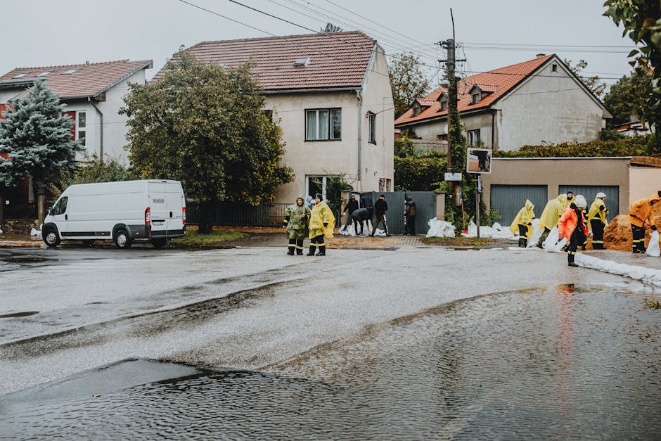 A group of emergency rescue personnel and civilians wading through a flooded street during a flood response. The rescuers are wearing bright orange life jackets and protective helmets, while some civi