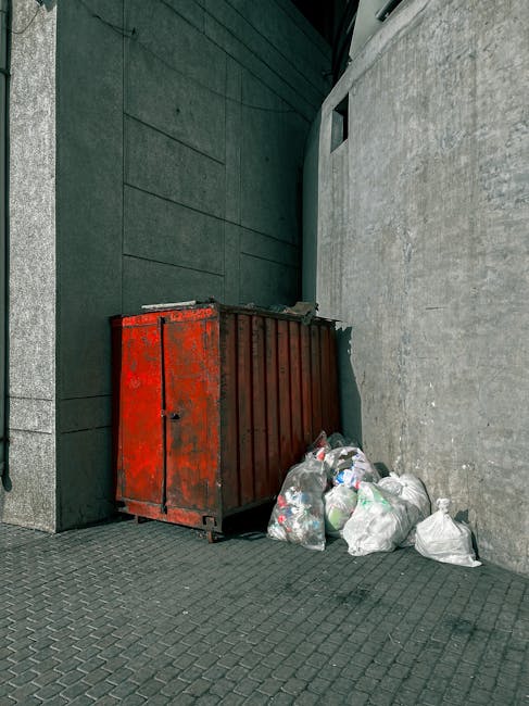 A close-up view of a cylindrical metal wire mesh bin filled with crumpled white paper trash, placed on a textured brown carpeted floor in an indoor space. The bin appears clean, with some scattered cr